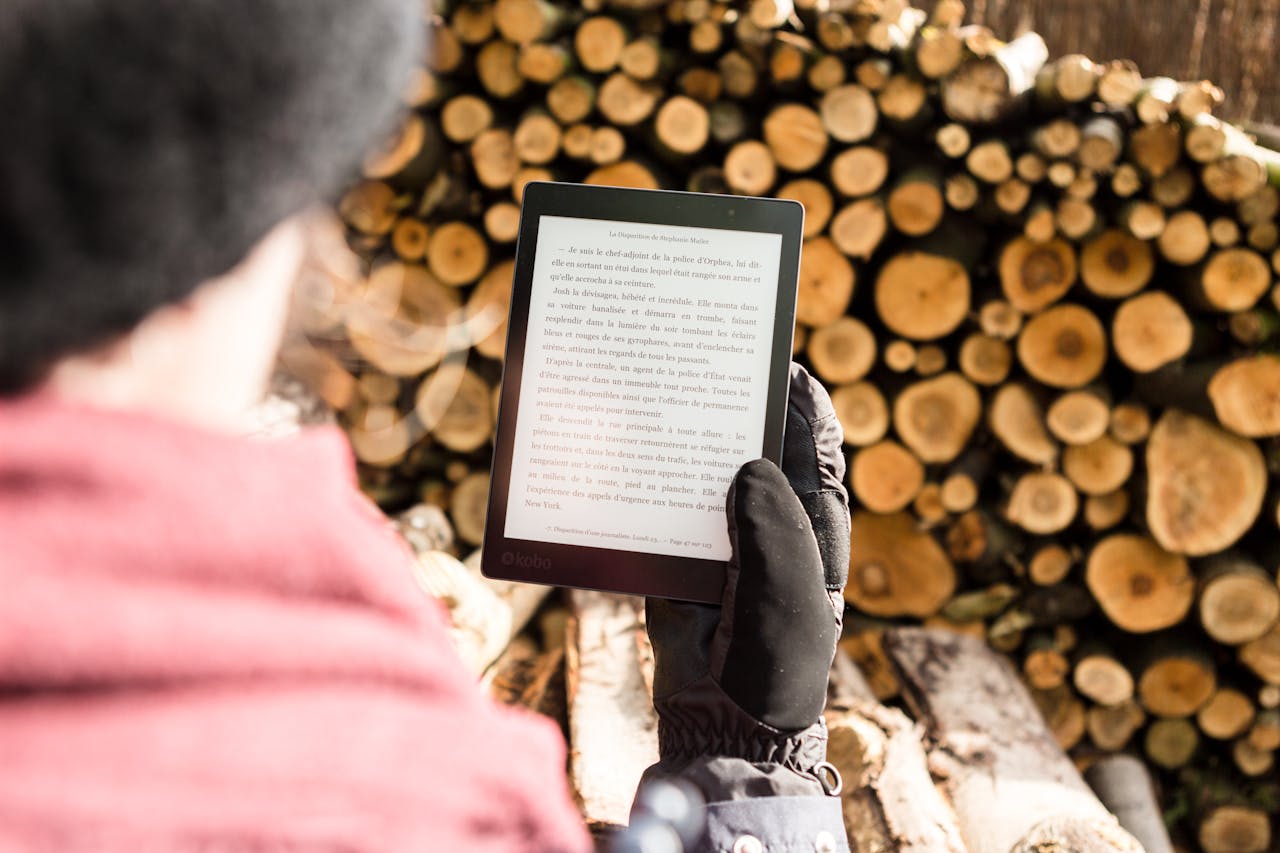 services-bg Person in warm clothing reads an e-book outdoors near a pile of firewood.